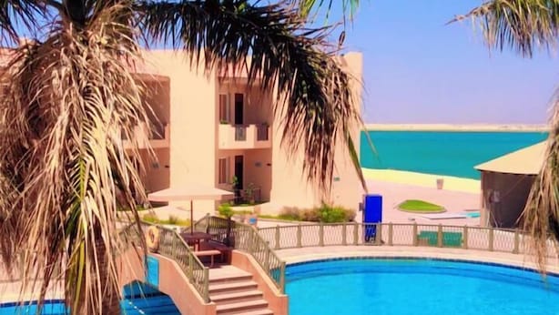 A swimming pool surrounded by palm trees and a building with a balcony, overlooking a beach. - ALAHLAM CHALET YANBU (Yanbu)