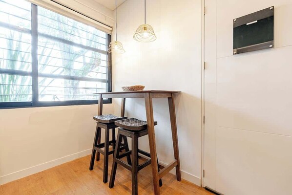 A corner room with a wooden floor, two stools, a small table with a basket, and a window with blinds.