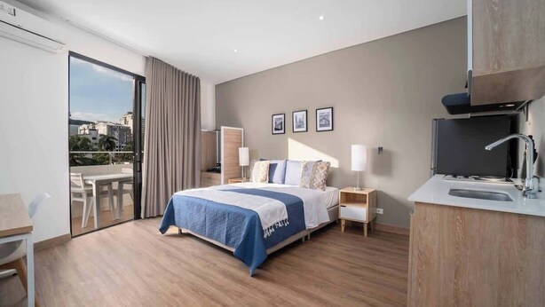 A modern hotel room with a large bed, a desk, a chair, a TV, and a sliding door to a balcony.