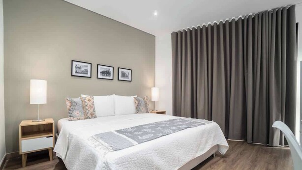 Une chambre d’hôtel moderne avec un grand lit, deux tables de chevet, une petite table et une fenêtre avec des rideaux.