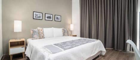 A modern hotel room with a large bed, two bedside tables, a nightstand, and a window with curtains.