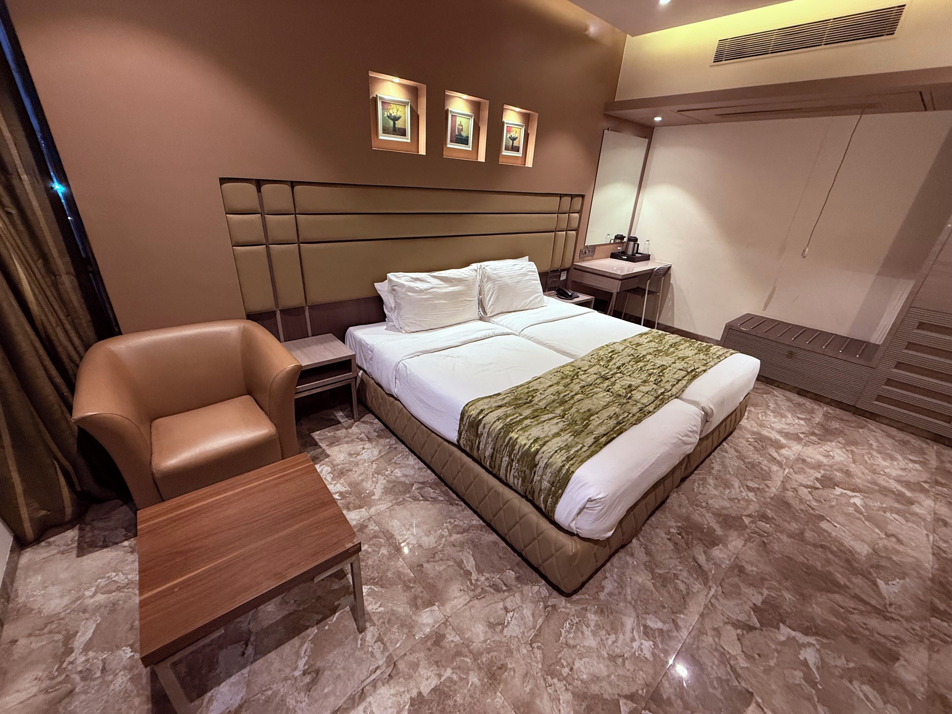A hotel room with a bed, a chair, a nightstand, a desk, and a wall-mounted shelf.