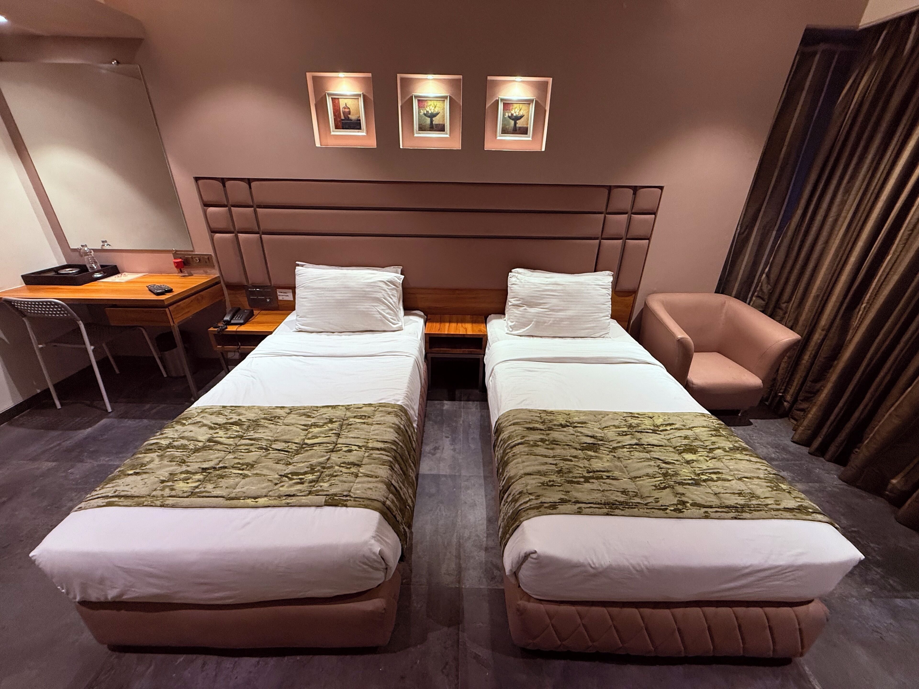 A hotel room with two beds, a desk, a chair, and three framed pictures on the wall.