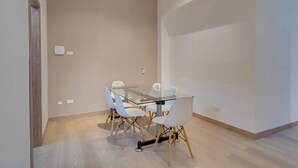 Una habitación moderna con una mesa de vidrio y sillas blancas, piso de madera y una pared lisa con un interruptor de luz.