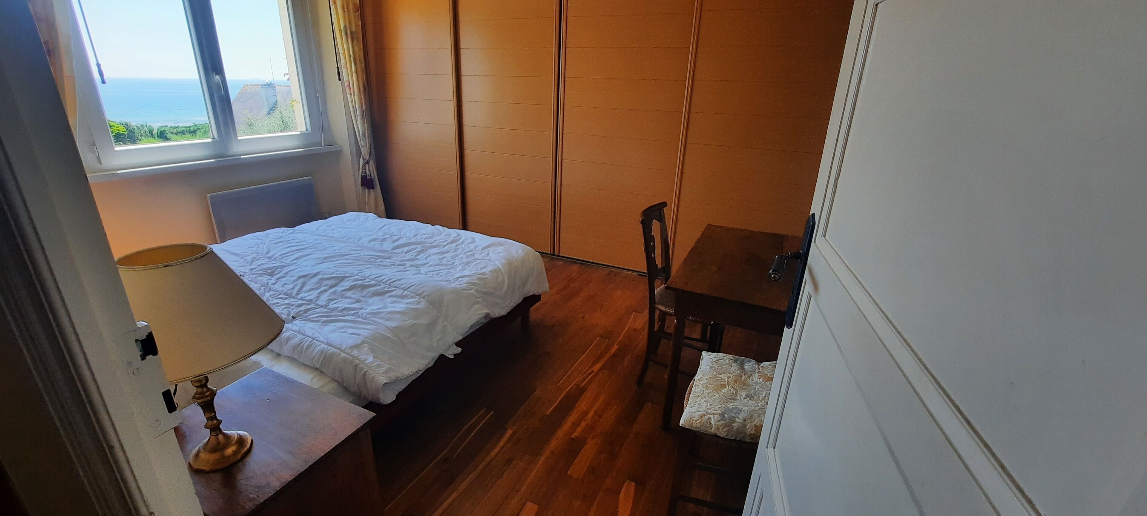 A bedroom with a bed, a chair, a table, and a window with a view of the sea.