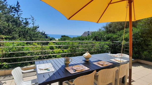 Una terraza con una mesa lista para dos, una sombrilla amarilla y vista al mar.