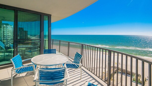 Ein Balkon mit rundem Tisch und blauen Stühlen, der auf einen Strand mit Meer und blauem Himmel blickt.