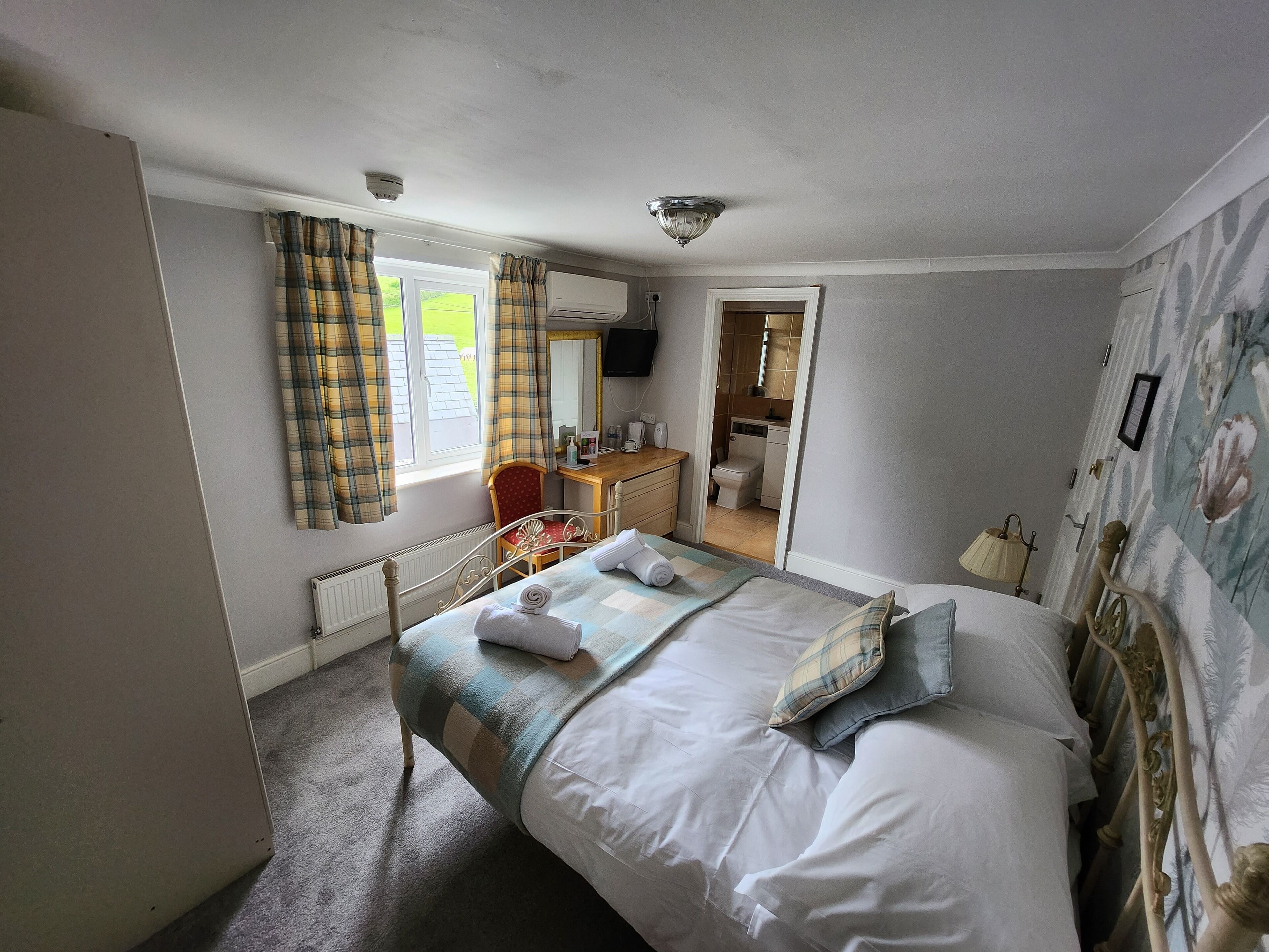 Une chambre à coucher comprenant un lit, un bureau, une chaise, une fenêtre avec des rideaux et une salle de bain visible à travers une porte ouverte.