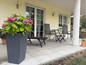 An outdoor patio with a seating area, a large planter with flowers, and a building with windows and a door.