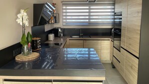 A modern kitchen with a black countertop, wooden cabinets, a coffee machine, and a window with blinds. - Superb villa with pool 10 minutes from the beach (Sérignan)