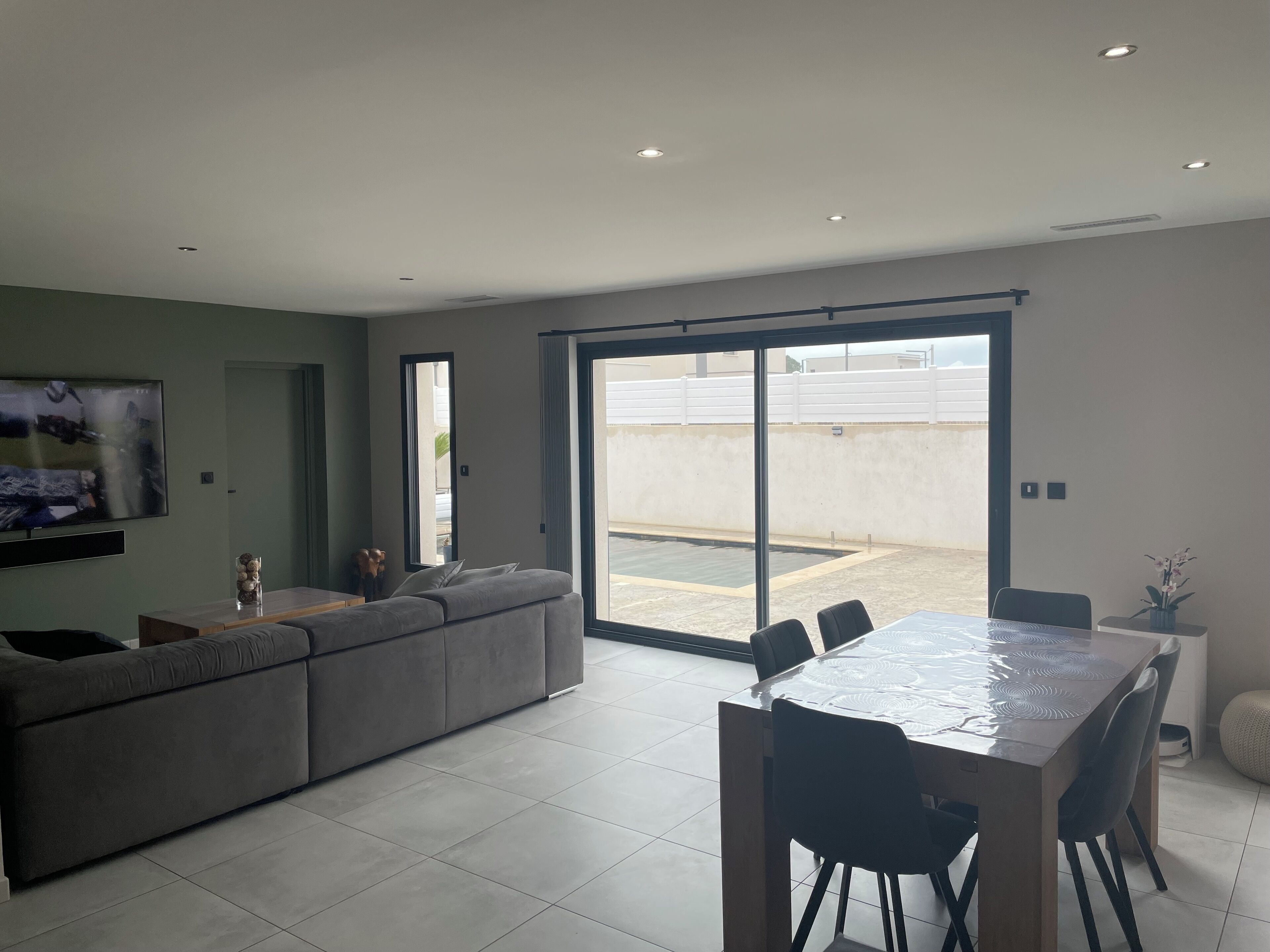 Una sala de estar moderna con un sofá seccional gris, una mesa de comedor con sillas, un televisor de pantalla plana montado en la pared y un cerramiento de vidrio corredizo que da a un patio exterior.
