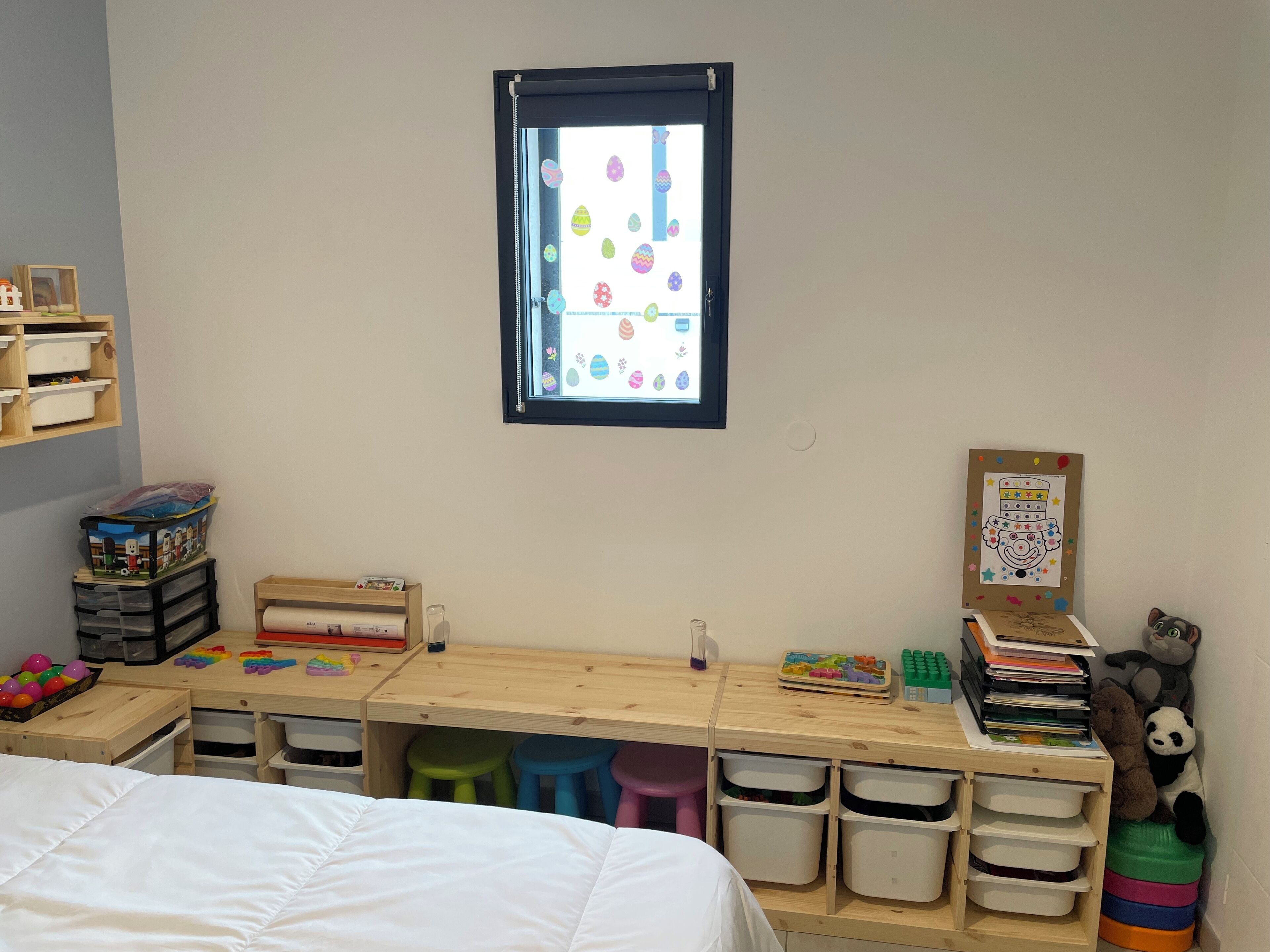 A child's room with a wooden desk, storage bins, a bed, and a window with a view of colorful Easter eggs.
