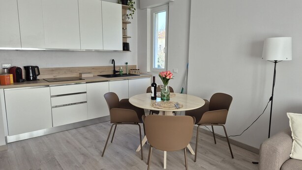 Una cucina moderna con mobili bianchi, un tavolo rotondo con sei sedie, una macchina per caffè e un vaso con fiori.