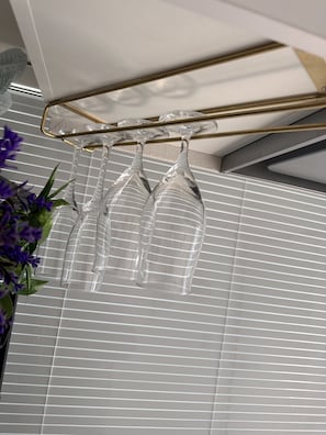 Wine glasses hang from a metal rack above a window blind, accompanied by a potted plant.