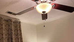 A bedroom with a bed, a nightstand with a lamp, patterned curtains, and a ceiling fan.