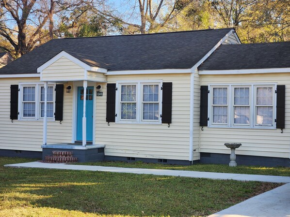 Una casa de un piso con puerta azul, revestimiento blanco y contraventanas negras.