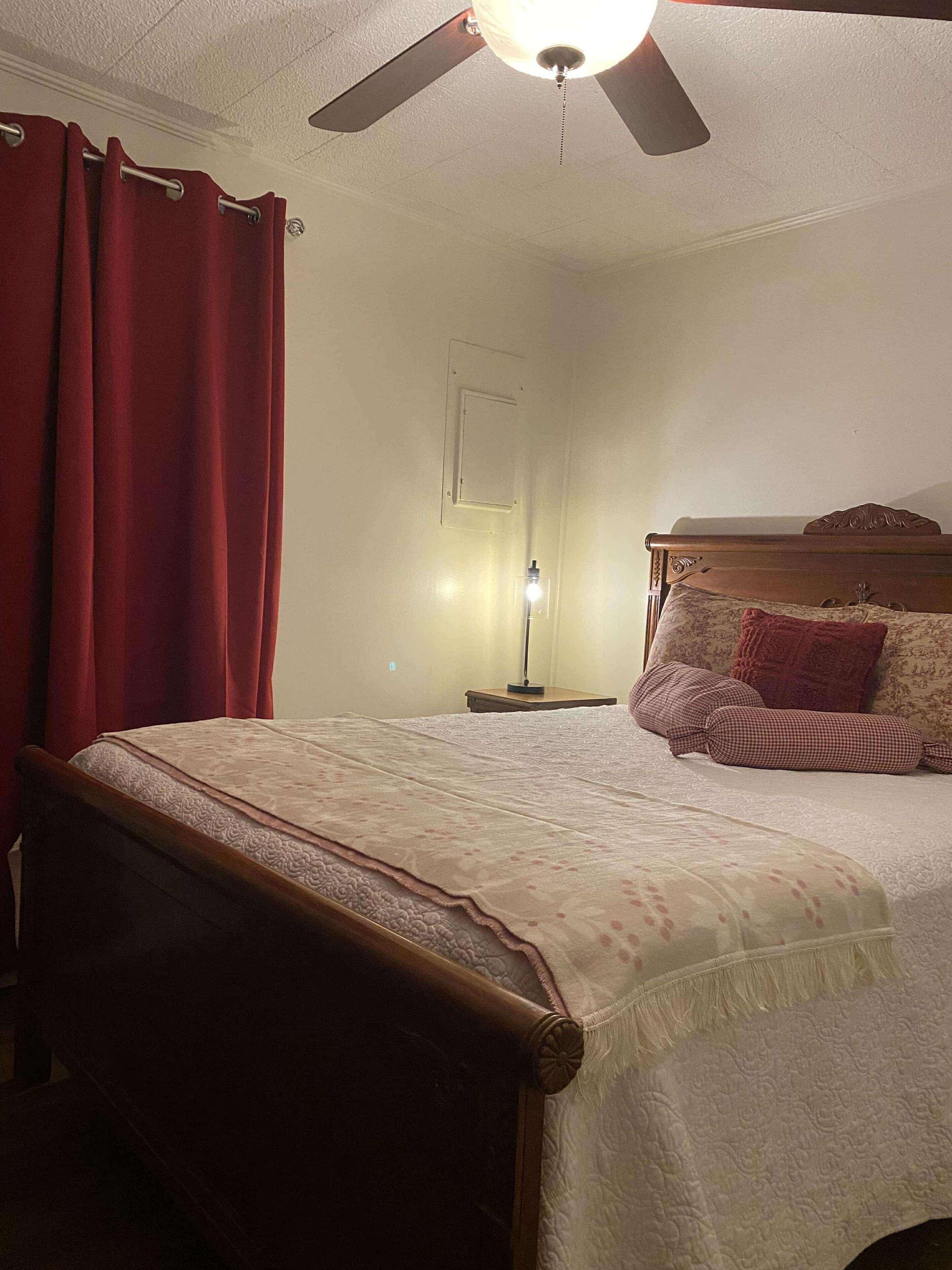 Een slaapkamer met een bed, een plafondventilator, een nachtkastje met lamp en een raam met rode gordijnen.