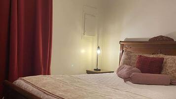 Een slaapkamer met een bed, een plafondventilator, een nachtkastje met lamp en een raam met rode gordijnen.