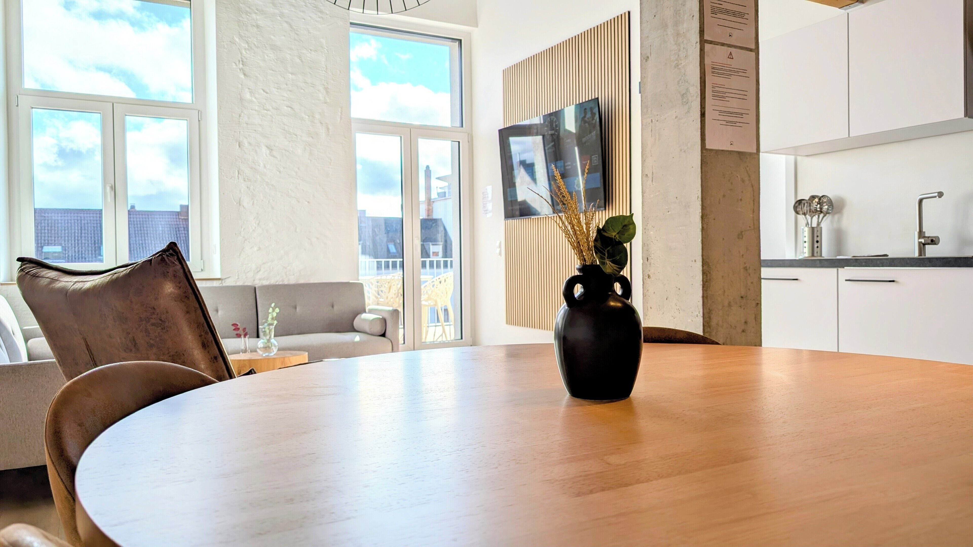 A modern living room with a wooden dining table, a black vase with dried plants, and a city view through large windows.