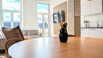 A modern living room with a wooden dining table, a black vase with dried plants, and a city view through large windows.