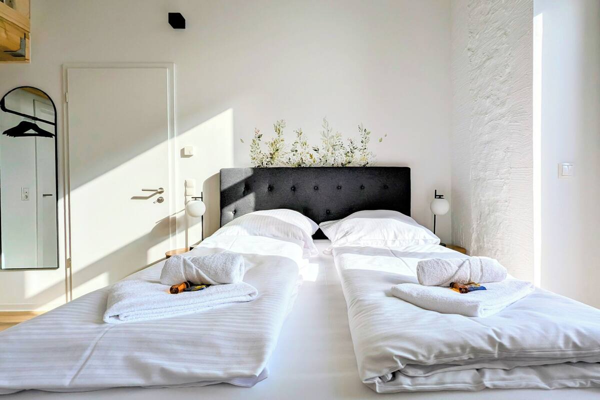 A double bed with white linens and a dark headboard, a bedside table with a lamp, and a mirror on the wall.