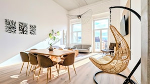Een moderne leefruimte met een houten eettafel, comfortabele zithoek, een hangstoel en een groot raam met uitzicht op de stad.