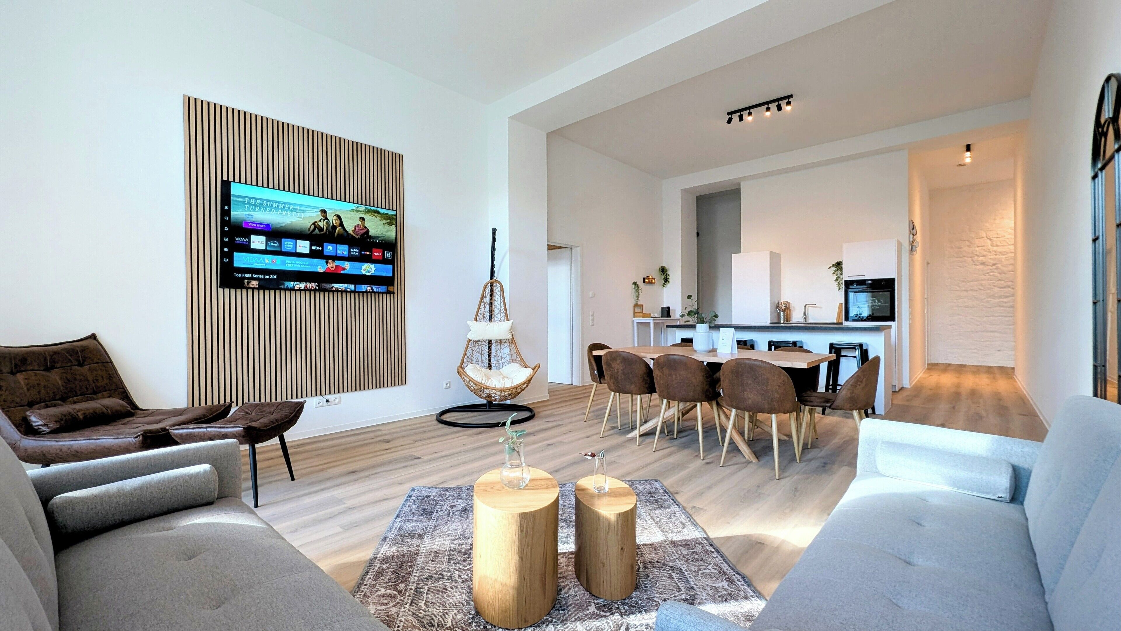 Una sala de estar moderna con un televisor de pantalla plana, un sillón colgante, un comedor con una mesa y sillas de madera, y una cocina al fondo.