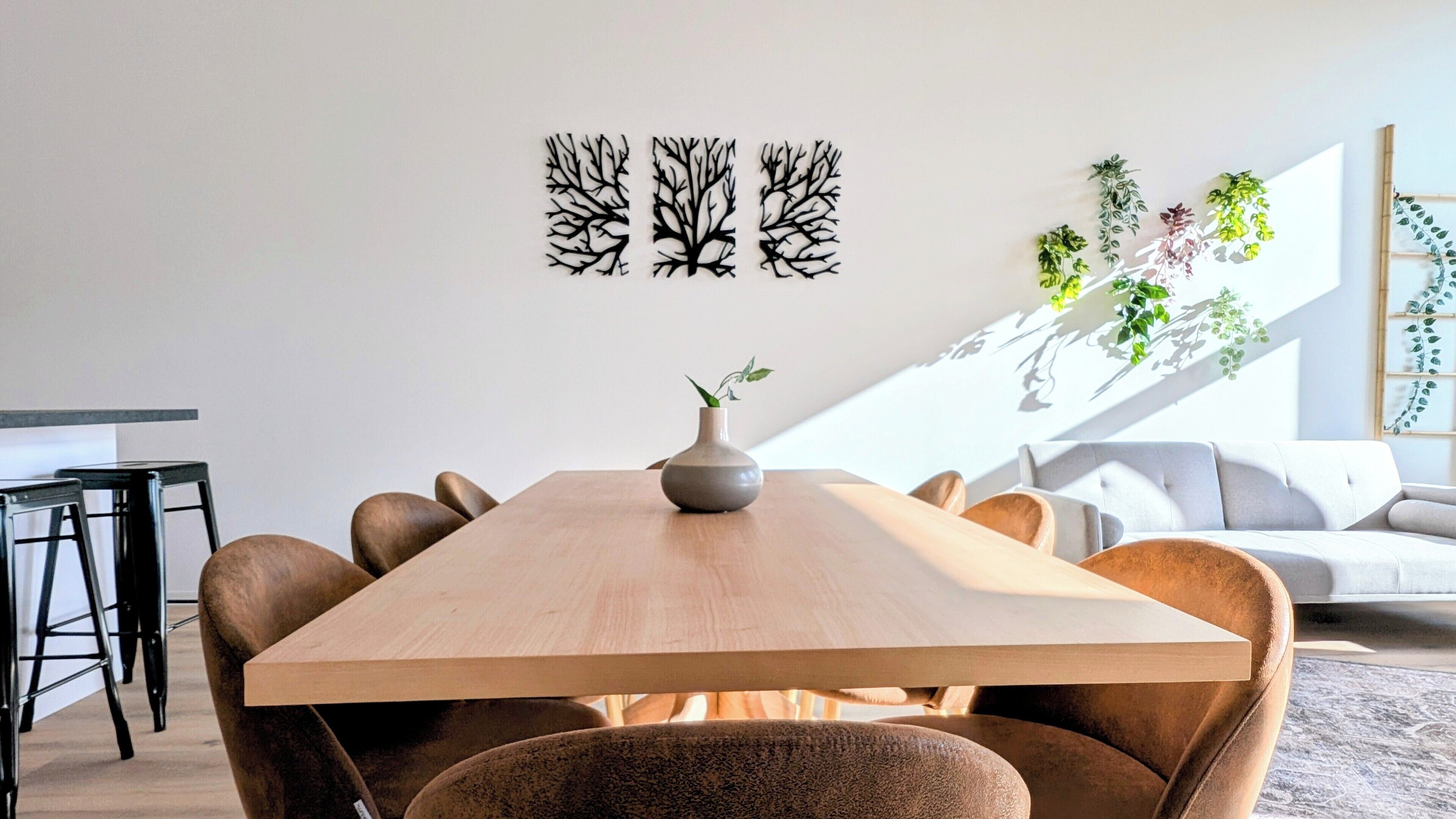 A modern dining area with a wooden table, chairs, and a vase with a plant. There are black wall art pieces above the table and potted plants on the wall.