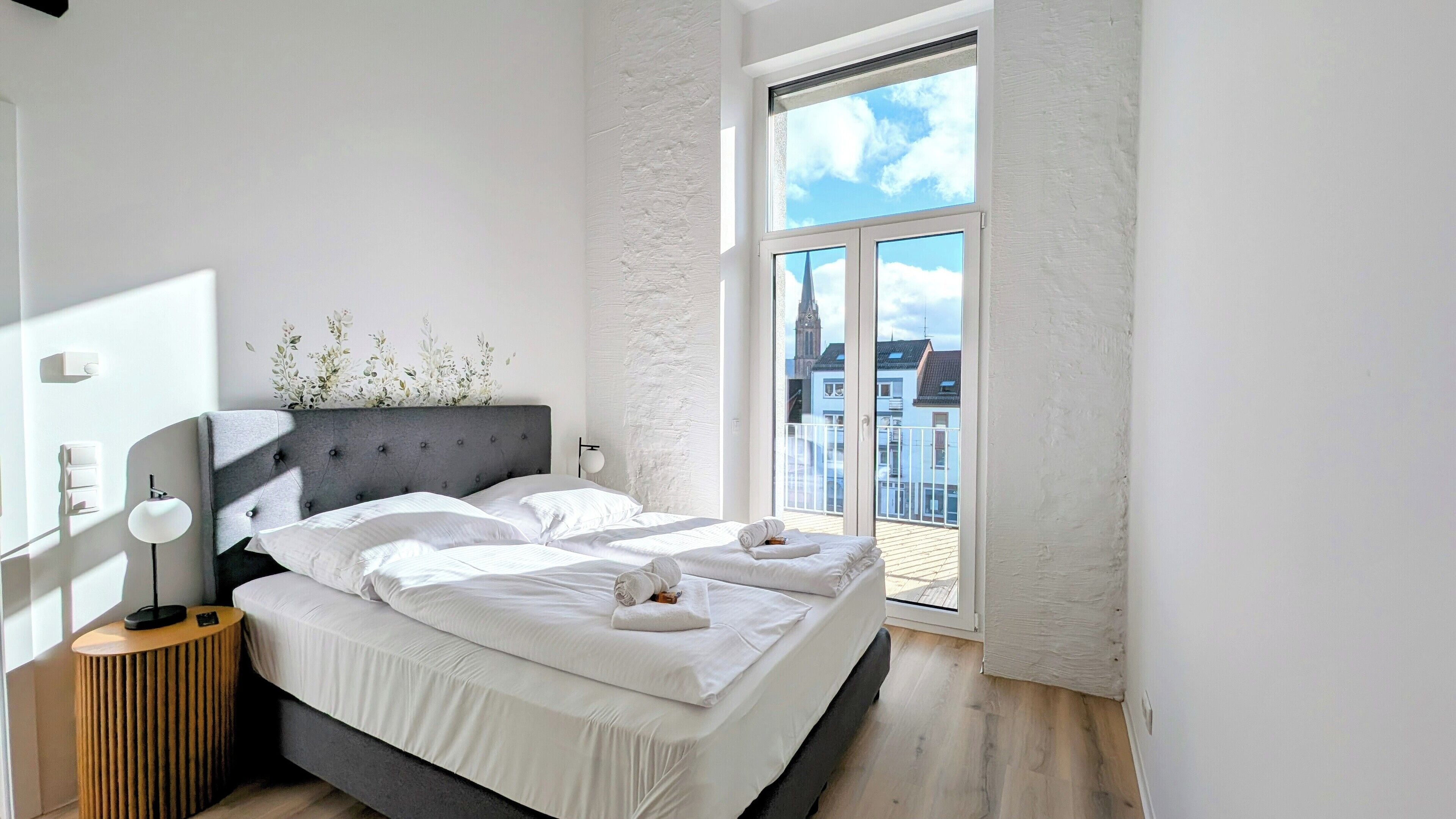A modern bedroom with a large bed, a bedside table, a lamp, and a view of buildings through a large window.