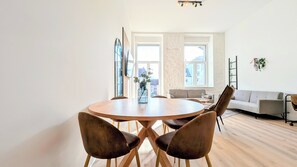 Un salon moderne avec une table à manger ronde en bois, deux chaises marron, un canapé gris et une grande fenêtre donnant sur des immeubles.