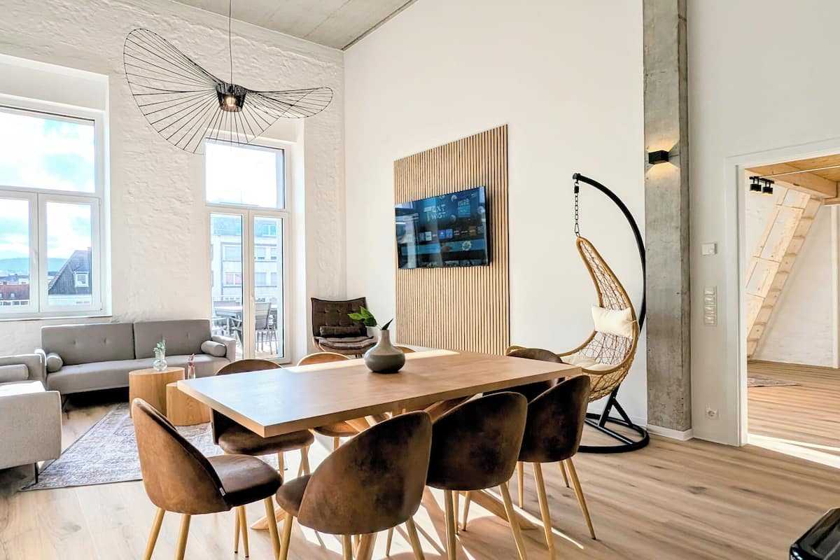 A modern living room with a wooden dining table, comfortable seating, and a large window offering a city view.