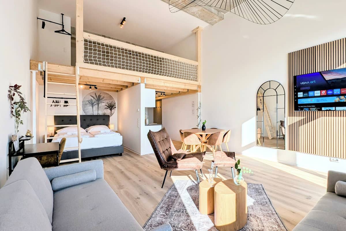 A modern living room with a loft bedroom, a TV mounted on a wooden panel wall, and a dining area with a table and chairs.