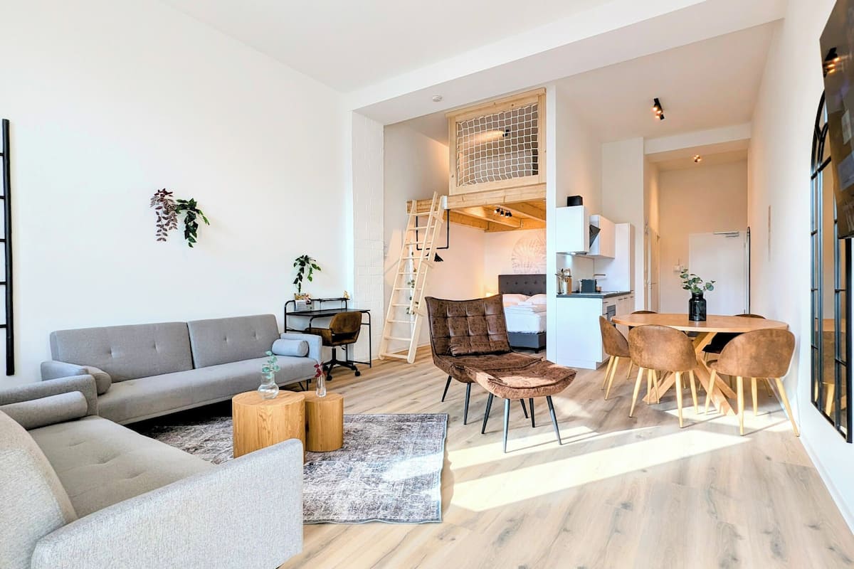 A modern living room with a grey sofa, a brown armchair, and a wooden coffee table. There is a dining area with a wooden table and chairs.