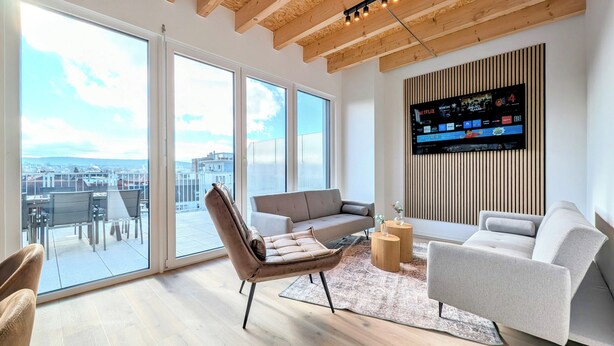 Een moderne woonkamer met een grote televisie, comfortabele zithoek en uitzicht op de stad door grote ramen.