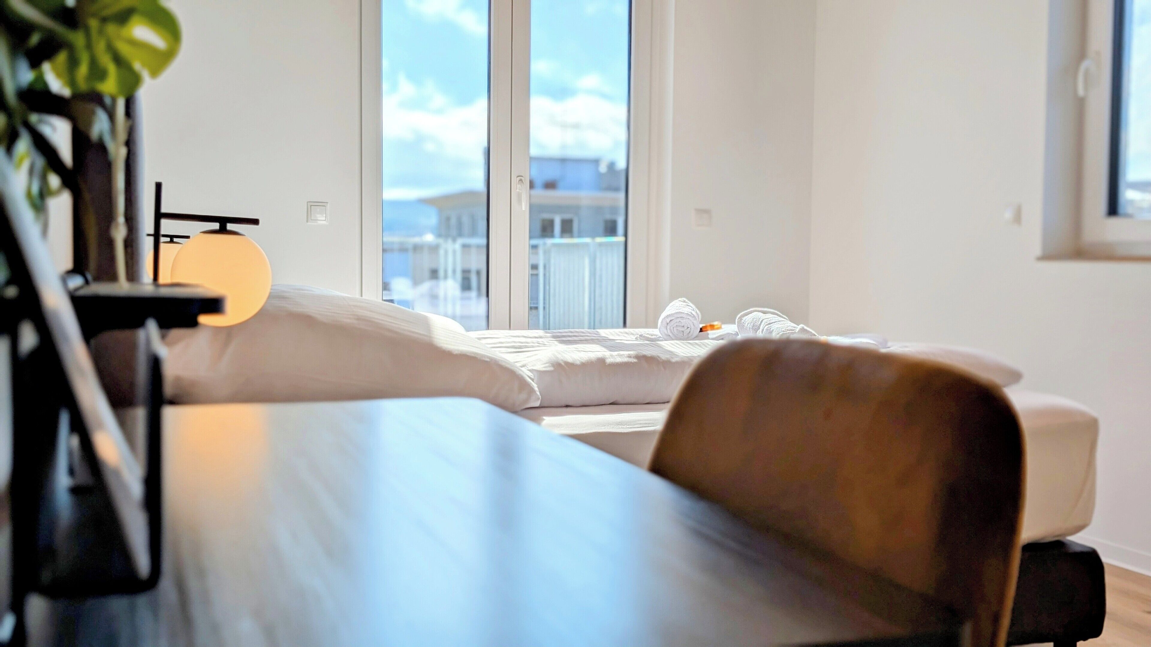 Moderni hotellihuone, jossa on suuri ikkuna, valkoisilla lakanoiden peitetty sänky, puinen työpöytä ja tuoli.