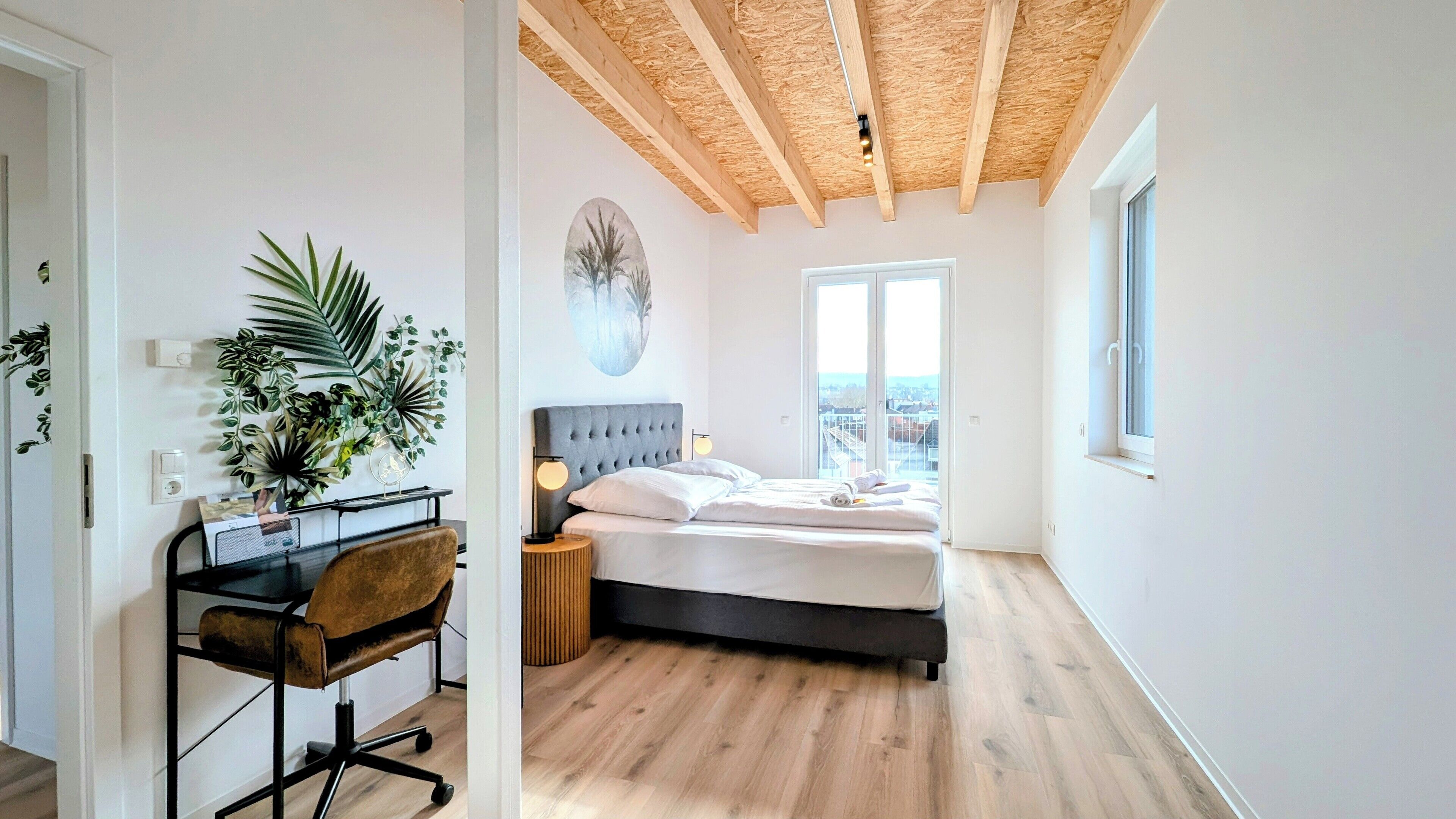 A bedroom with a wooden ceiling, a bed with white bedding, a desk with a chair, and a large window offering a city view.