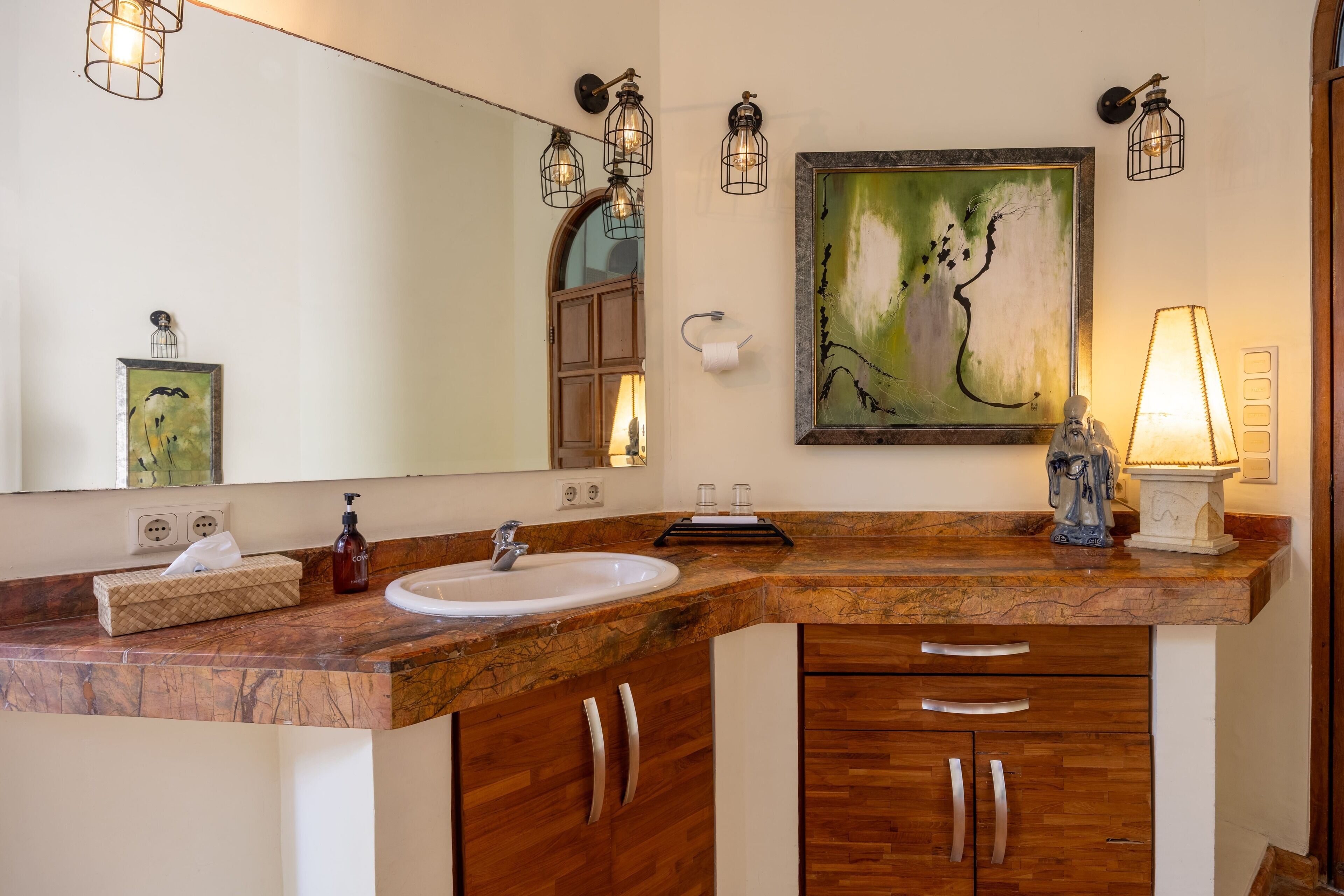 Une salle de bain avec un comptoir en marbre, des meubles en bois, un lavabo, un miroir, un tableau et des appliques murales.