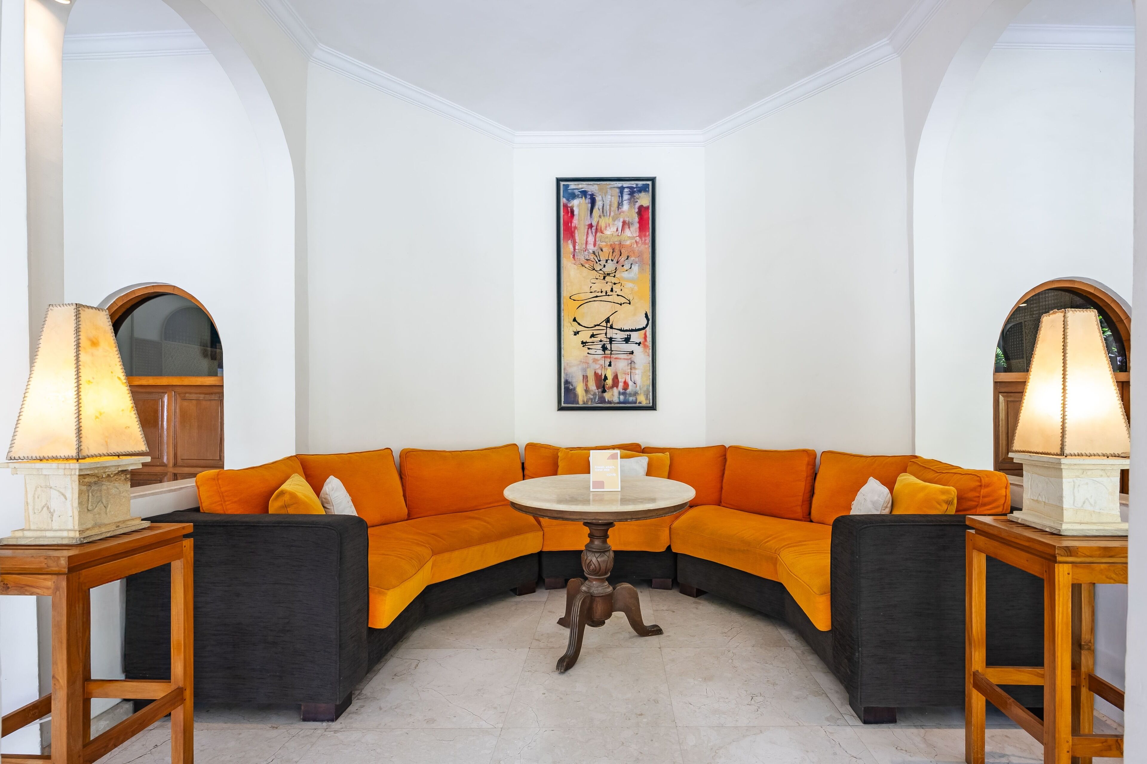 A modern living room with a curved orange sofa, a round wooden table, and two lamps on wooden stands. A colorful abstract painting is on the wall above the sofa.