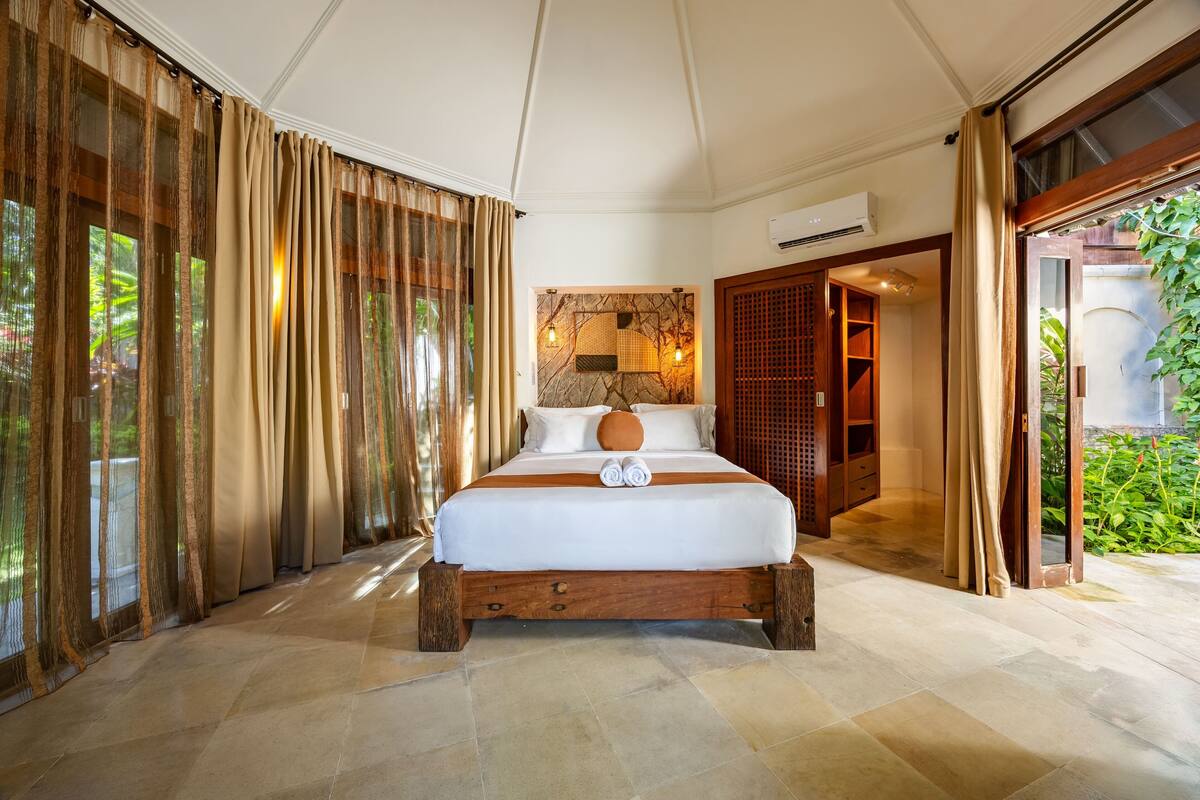 Une chambre spacieuse avec un grand lit, une tête de lit en bois et une vue sur la verdure environnante.