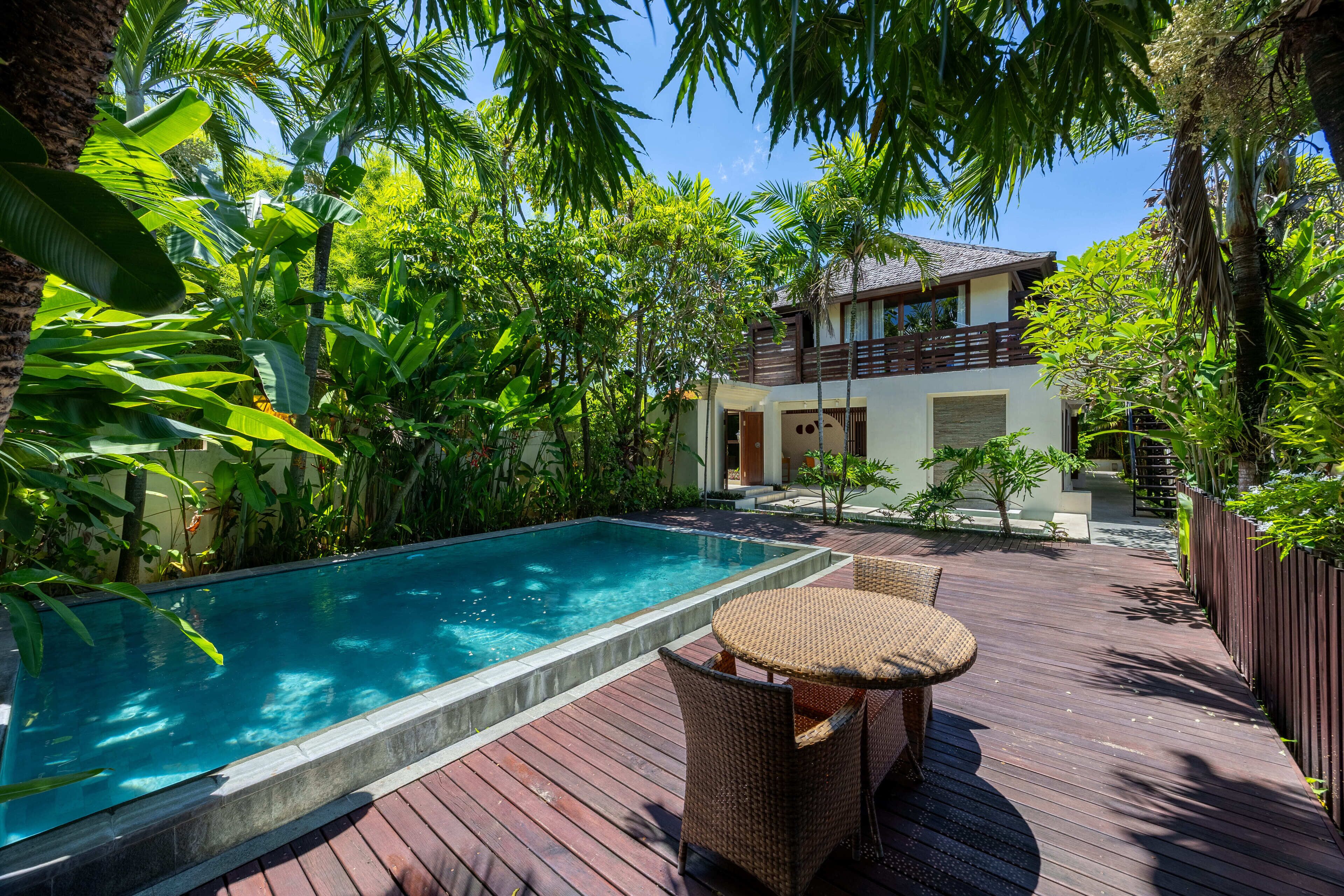 Una piscina rodeada de exuberante vegetación, una terraza de madera y una casa de dos pisos con balcón.