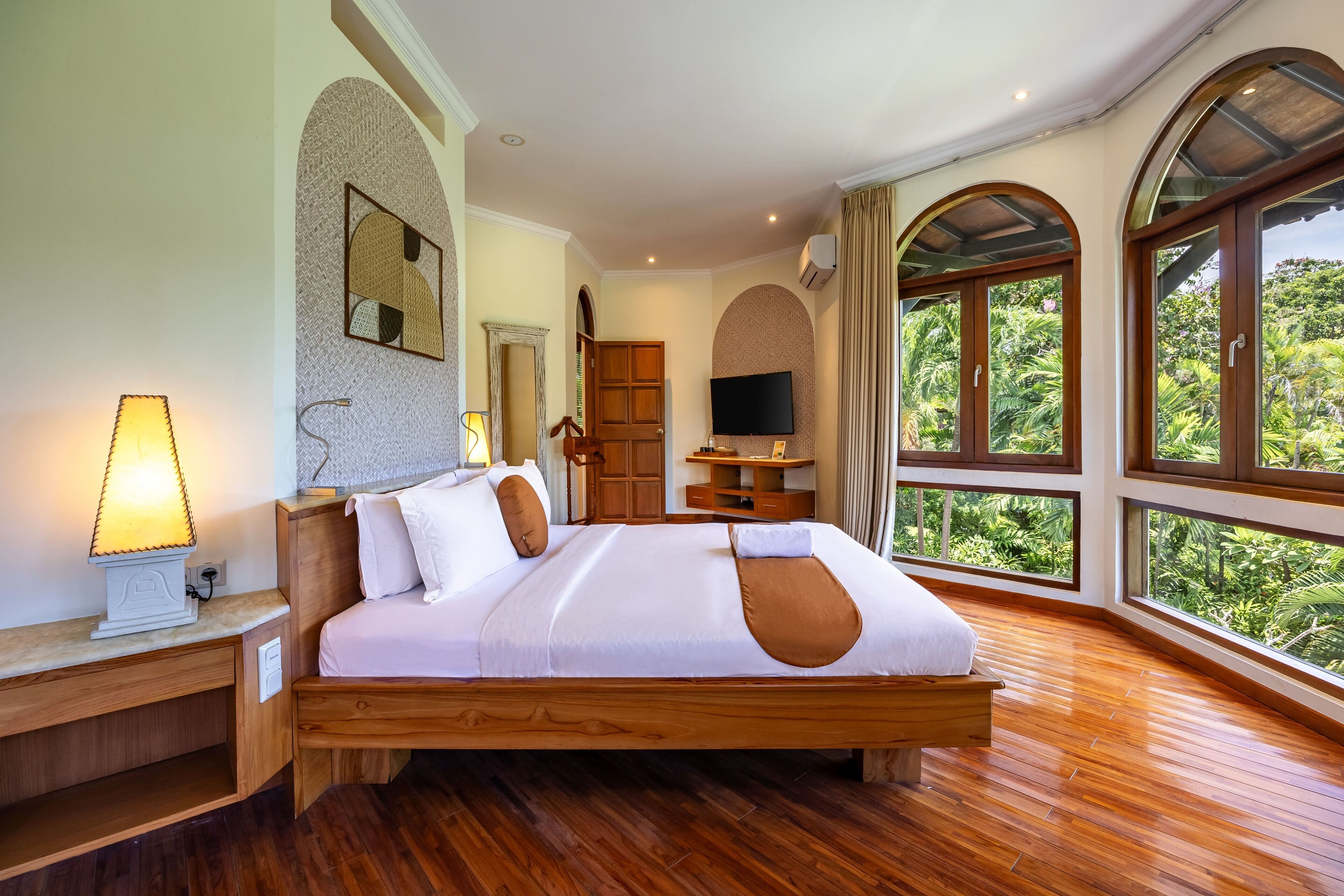 A spacious bedroom with a large bed, wooden flooring, a TV, and large windows offering a view of greenery.