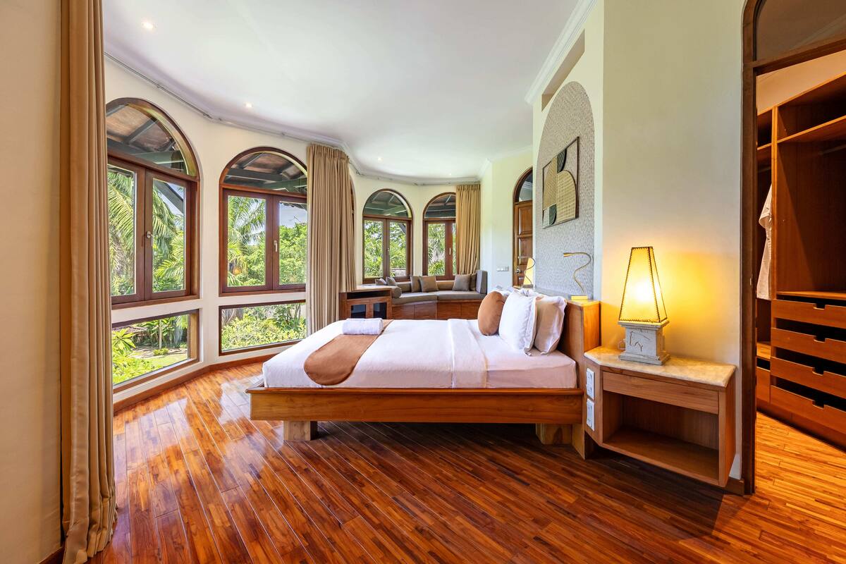 Une chambre spacieuse avec un grand lit, un plancher en bois et de nombreuses fenêtres donnant sur un paysage verdoyant.