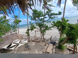 A beach view with a thatched-roof structure, wooden tables, and chairs, surrounded by palm trees and a clear sky. - Kamp Tapik - Sibaltan (El Nido)