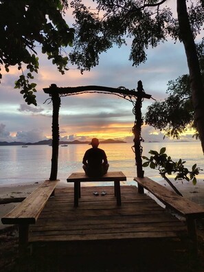 A wooden deck with a bench and a table, overlooking a body of water with boats and distant land. - Kamp Tapik - Sibaltan (El Nido)