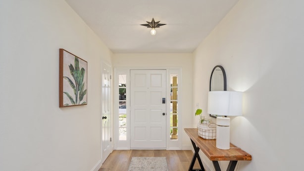 A hallway with a wooden floor, a white door, a framed plant, and a table with a lamp and decorative items.