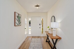 A hallway with a wooden floor, a white door, a framed plant, and a table with a lamp and decorative items.
