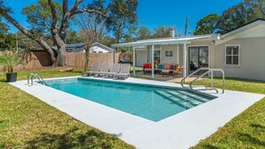 A rectangular swimming pool with a white deck, surrounded by a grassy area. There are lounge chairs and a covered patio area with a sofa and cushions.