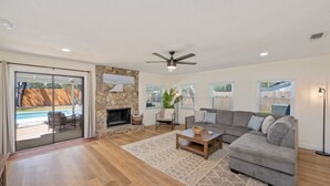 A spacious living room with a grey sectional sofa, a wooden coffee table, and a stone fireplace. There is a large sliding glass door leading to an outdoor area with a pool.