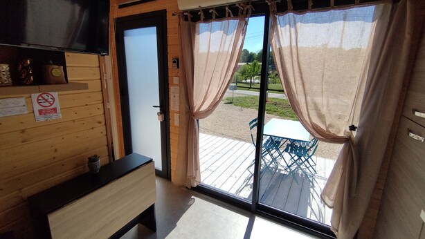 Une pièce au décor intérieur en bois, avec une porte-fenêtre coulissante donnant sur un espace extérieur équipé d’une table et de chaises, et un téléviseur fixé au mur.
