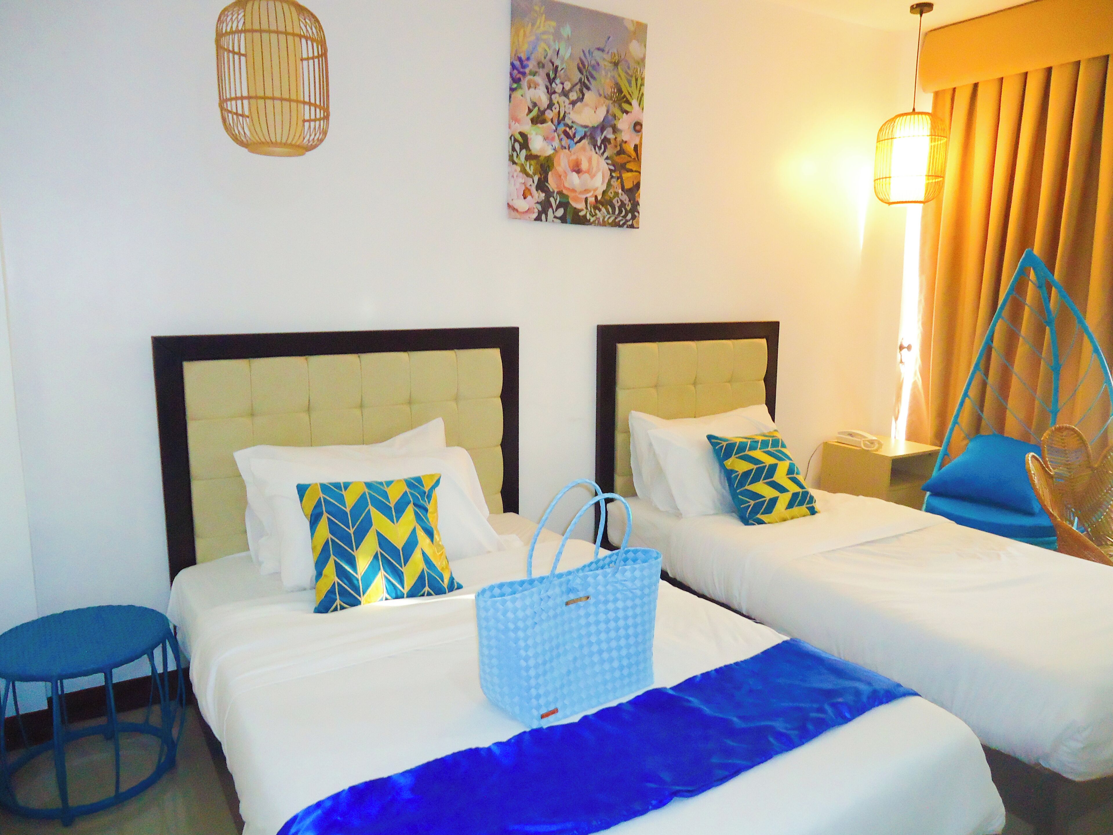 Een hotelkamer met twee bedden, een blauwe stoel, een rieten stoel, een blauwe tas en een schilderij aan de muur.